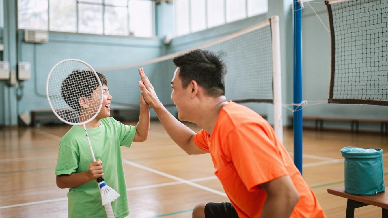 A Malay Malaysian father in an orange t-shirt giving a high-five to his young son holding a badminton racket and shuttlecock on an indoor badminton court