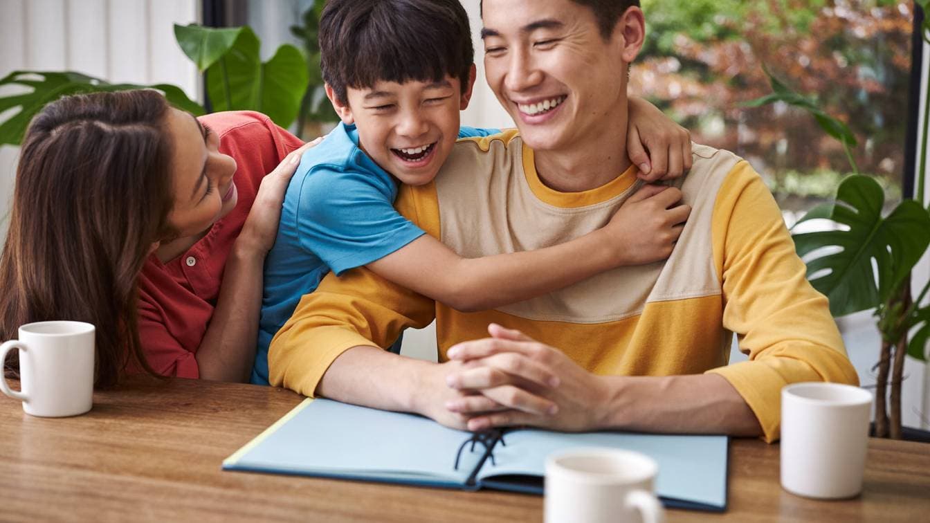 A laughing Chinese Malaysian boy hugging his father at a sunny kitchen table while his mother leans in smiling, with an open blank notebook and white mugs on the table