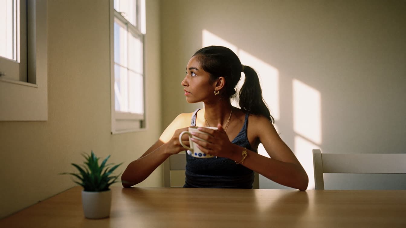 A young woman sitting with a mug at a bare wooden table in a bright empty room beside a sunlit window