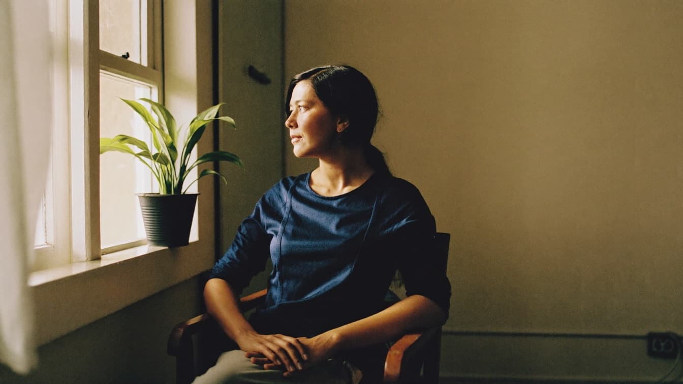 A woman sitting quietly beside a sunlit window in a bare room with a single houseplant
