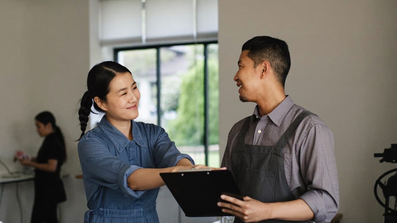 Two Malaysian small-business partners discussing work over a clipboard in their bright workshop