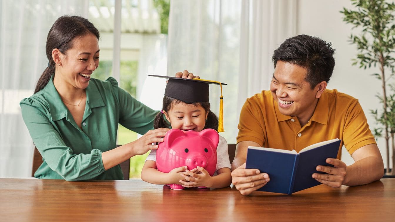 A Chinese Malaysian couple laughing with their young daughter, who wears a graduation cap and hugs a pink piggy bank at the dining table